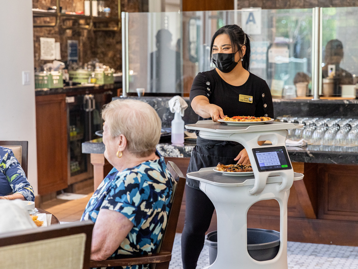 A staff member at La Costa Glen senior center smiles at residents as they serve their food with the help of Servi, Bear Robotics' autonomous robot waiter.